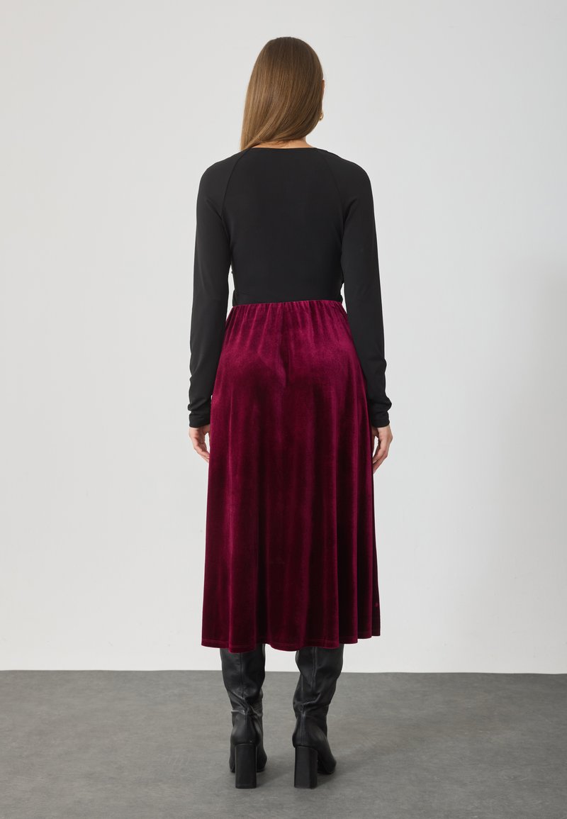 Top negru cu mânecă lungă și guler rotund, asortat cu o fustă de catifea burgundă, de lungime medie. Costumul are o talie strânsă și o textură netedă.