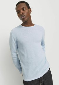 Jersey azul claro de manga larga con un diseño de punto texturizado y cuello redondo, combinado con jeans oscuros. Corte simple y moderno.