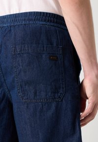 Tmavě modré džínové šortky s elastickým pasem, které mají zadní kapsu s malou hnědou etikettou. Hladká textura s detaily šití.