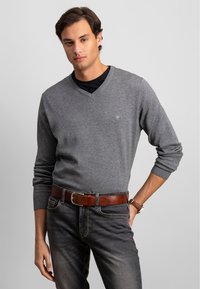 Grå V-hals genser laget av myk stoff, lange ermer, ribbede mansjetter, paret med mørke jeans og et brunt lærbelte. Ingen synlige mønstre.