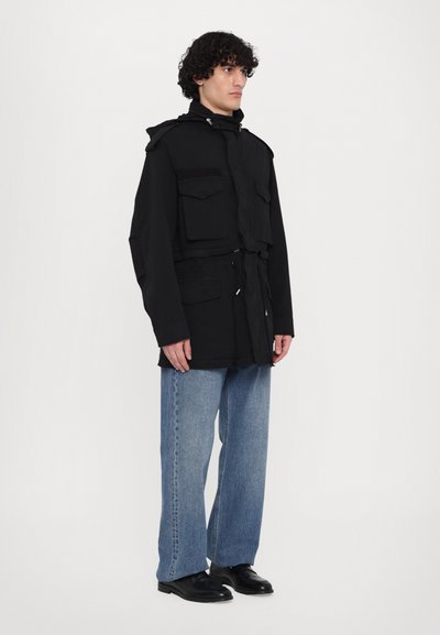 Zwarte jas met een trekkoord in de taille, twee borstzakken en een opstaande kraag, gedragen met lichtblauwe wijde jeans en zwarte schoenen.