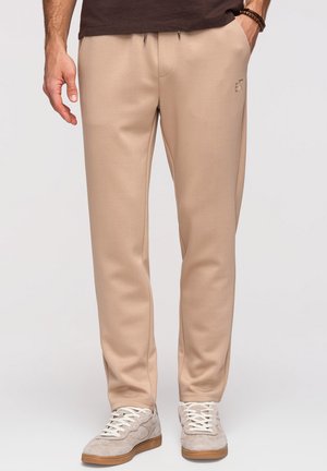 Homme portant un pantalon de jogging beige avec le logo "EX" et des baskets gris clair, debout devant un fond gris uni.