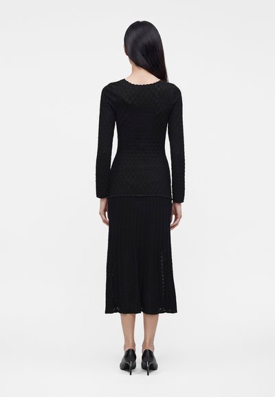 Femme aux longs cheveux noirs portant un haut noir texturé à manches longues, une jupe midi noire et des chaussures à talons noirs, vue de dos.