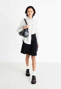 Chemise à rayures, jupe noire évasée, sac bandoulière noir, chaussures sombres épaisses et chaussettes blanches. Tenue simple et polyvalente.