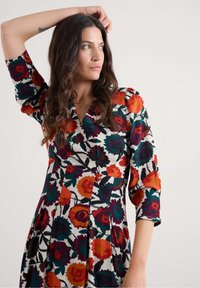 Žena s dugom valovitom kosom koja nosi šarene cvjetne haljine, podiže lijevu ruku i gleda sa strane protiv jednostavne pozadine.