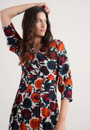 Femme aux longs cheveux ondulés portant une robe florale colorée, levant le bras gauche et regardant sur le côté devant un fond uni.