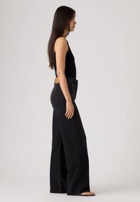 Γυναίκα με μακριά σκούρα μαλλιά, που φοράει μαύρο αμάνικο τοπ και ψηλόμεσα μαύρα τζιν με φαρδιά πόδια, όρθια ξυπόλυτη σε πλάγια όψη.