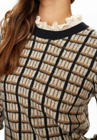 Pull en maille présentant un motif quadrillé en crème et marron, avec des accents noirs. Le col à volants ajoute de la texture. Gros plan sur l'encolure et le tissu.