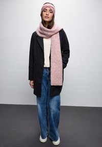 Cappotto nero, maglione bianco a trecce, jeans blu larghi, sciarpa rosa e fascia per capelli. La sciarpa ha un motivo sottile. La texture della lana pettinata è evidente.