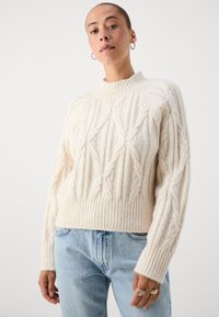 Κρεμ πλεκτή μπλούζα με ψηλό ριμπ λαιμό κ αι διαμάντια από καμπύλες. Διαθέτει ριμπ μανσέτες και στρίφωμα, συνδυασμένη με ανοιχτό μπλε τζιν.