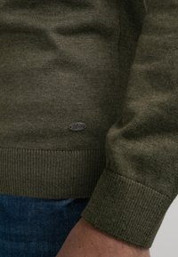 Olijfgroene trui in gebreide textuur met geribde manchetten en zoom. Heeft een klein logotag bij de zoom. G gedragen over spijkerbroeken.