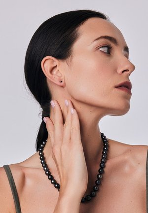 Czarna perłowa naszyjnik na nagim ramieniu, gładka tekstura. Modelka ma rękę na szyi, nosi małe kolczyki wkręty, lekko pomalowane paznokcie.