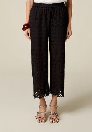 Zwarte broek met wijde pijpen met bloemmotiefuitsparingen en geschulpte zomen, gedragen met sandaaltjes met slangenprint en bruine armbanden om de linker pols.