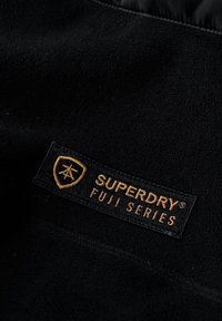 Zwarte fleece stof met een gestructureerd oppervlak, met een rechthoekige patch voorzien van de tekst "SUPERDRY FUJI SERIES" in gouden letters op een zwarte achtergrond.