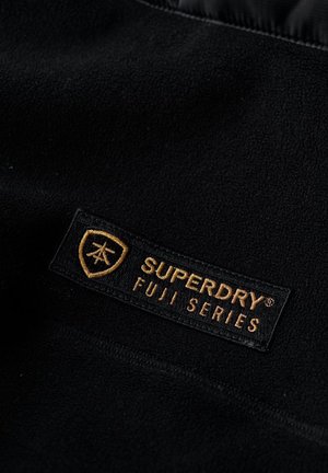 Zwarte fleece stof met een gestructureerd oppervlak, met een rechthoekige patch voorzien van de tekst "SUPERDRY FUJI SERIES" in gouden letters op een zwarte achtergrond.