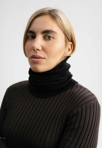 Zwarte geribde coltrui met een hoge kraag, voorzien van verticale strepen en een gestructureerd breipatroon, gedragen over een donkerbruine langwerpige top.