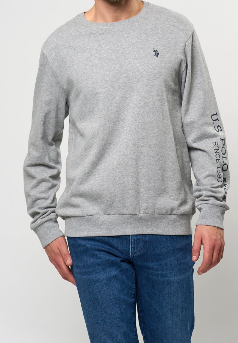 Homme portant un sweat-shirt gris à manches longues avec un petit logo sur la poitrine et le texte "U.S. Polo" sur la manche droite, associé à un jean bleu.