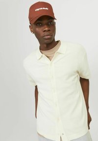 Lühikeste varrukatega kreemika värvusega nööbid-särk, millel on tekstuuriga kangas ja klassikaline krae, koos tumeda pruunist mütsiga, millel on valged kirjad.