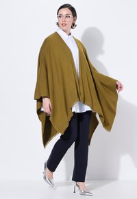 Olivgrüner Poncho mit Fransen und durchbrochener Verzierung, getragen über einem weißen Hemd und einer marineblauen Hose; kombiniert mit silbernen Absätzen.