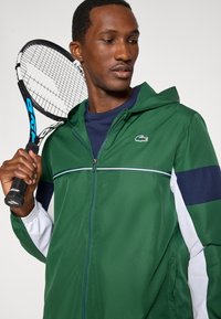 Zielona sportowa kurtka z białymi i granatowymi akcentami, zapinana na zamek błyskawiczny z przodu, z kapturem i logo na piersi; osoba trzymająca rakietę tenisową.