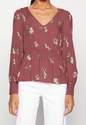 Kvinde iført en langærmet, V-udskåret, støvet rosa bluse med blomster- og sommerfugleprint, kombineret med hvide bukser.