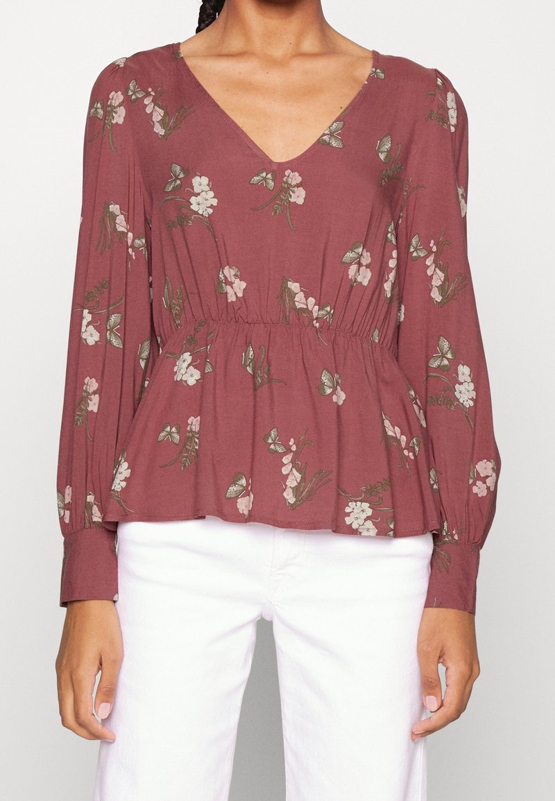 Femme portant un chemisier rose poudré à manches longues, col en V, avec un imprimé floral et papillons, associé à un pantalon blanc.