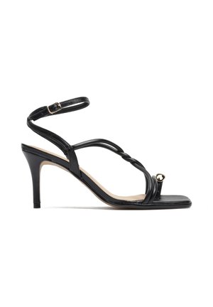 Czarna sandałka na wysokim obcasie z paskiem wokół kostki, skręcanymi przednimi paskami oraz małym złotym serduszkiem na pętli przy palcach.