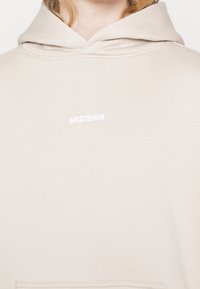 Sweat à capuche beige avec un petit logo blanc "WRSTBHVR" centré sur la poitrine et une capuche visible autour du cou.