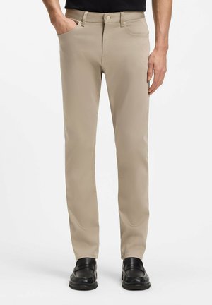 Uomo che indossa pantaloni beige a gamba dritta con tasche anteriori e mocassini neri, in piedi contro uno sfondo bianco semplice.