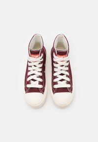 Wysokie trampki w kolorze bordowym z płótna, z białymi sznurówkami i gumowymi podeszwami. Posiadają pomarańczowe logo na języku oraz białe akcenty w strefach szwów.