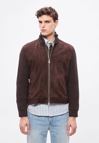 Jachetă bomber maro din piele întoarsă cu mâneci din tricot cu nervuri, închidere cu fermoar, buzunare laterale și guler înalt, purtată peste o cămașă în dungi.