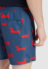 Costumi da bagno in tessuto blu navy con stampa di bassotti rossi. Presentano un elastico in vita e una tasca posteriore con patta. Texture liscia.