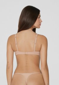 Žena s dlouhými tmavými vlasy, oblečena v nahých podprsenkových a thong kalhotkách, zobrazená zezadu na jednolitém světle šedém pozadí.