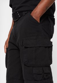 Mężczyzna w czarnych szortach cargo z wieloma kieszeniami i czarnym wplecionym paskiem, z ręką swobodnie w przedniej kieszeni.