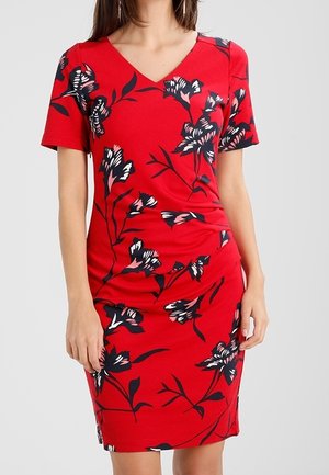 Kobieta w czerwonej sukience za kolano z czarno-białym kwiatowym wzorem i krótkimi rękawami na jasnym, jednolitym tle.