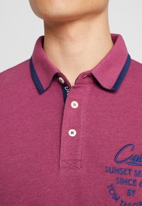 Burgundy piké med marinblå krage, tre knappar och broderad detalj. Tillverkad av texturerad tyg, med synliga sömnadsdetaljer.