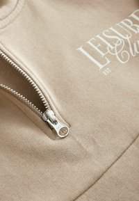 Beige zip-up sweatshirt med framträdande vit text "LEISURE CLUB". Texturerat tyg och silverfärgad dragkedjedetalj förbättrar designen.