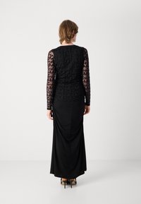 Schwarzes Kleid mit langen, durchsichtigen Ärmeln und einem gemusterten Überzug sowie einer figurbetonten Silhouette. Der Stoff ist glatt und leicht hinten drapiert.
