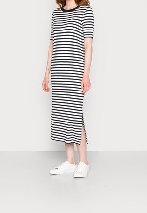 Mujer con un vestido midi de rayas horizontales en blanco y negro, de manga corta, y zapatillas blancas, de pie contra un fondo claro y liso.