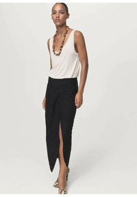 Falda larga negra con una abertura lateral, combinada con una blusa sin mangas beige. Un collar notable de cuentas y unos zapatos de diseño aportan un toque especial al conjunto.