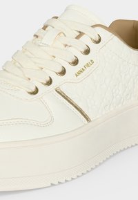 Close-up af hvid sneaker med hvide snørebånd, gyldne øjer, tekstureret sidepanel og guld "ANNA FIELD" logo over en gylden stribe.