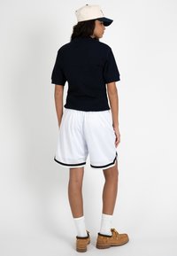 Personne vue de dos portant une casquette blanche, un t-shirt noir à manches courtes, un short de basket blanc avec des bordures noires, des chaussettes blanches et des chaussures marron.