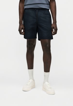 Άντρας που φοράει μαύρα σορτς με κορδόνι, λευκές κάλτσες και λευκά παπούτσια πλατφόρμας, στέκεται μπροστά σε απλό λευκό φόντο.