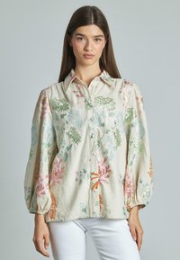 Blusa in lino beige con ricami floreali, dotata di maniche a sbuffo, chiusura frontale con bottoni e vivaci motivi floreali nei colori rosa, verde e blu.