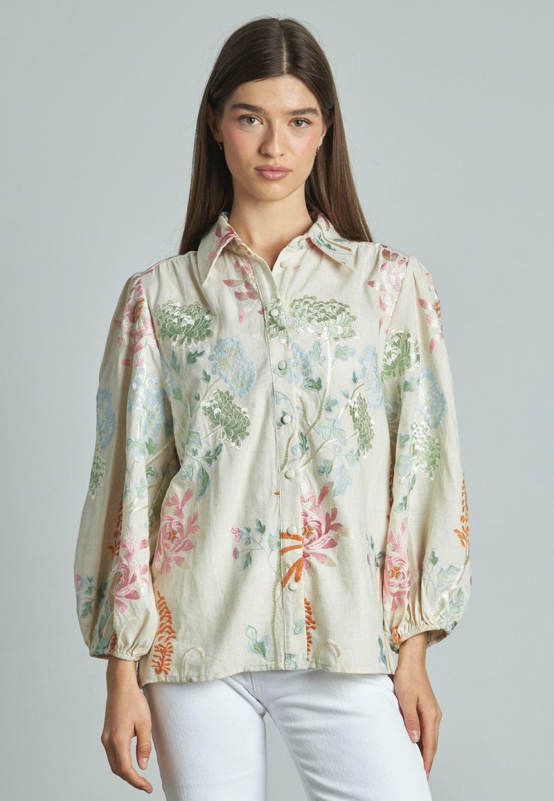 Blusa in lino beige con ricami floreali, dotata di maniche a sbuffo, chiusura frontale con bottoni e vivaci motivi floreali nei colori rosa, verde e blu.