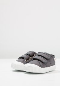 Scarpe in tela grigia con due cinturini regolabili in velcro e suole in gomma bianca. Materiale testurizzato e forma affusolata per il comfort.