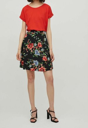 Haut rouge à manches courtes associé à une jupe noire à fleurs avec des fleurs rouges et bleues. Le mannequin porte des talons noirs à lanières.