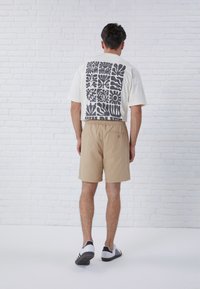 T-shirt bianco con design floreale astratto grigio sulla schiena, abbinato a pantaloncini beige. I pantaloncini hanno una cintura elastica e una tasca sul retro.