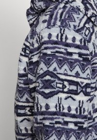 Giacca in fleece con un motivo geometrico viola e bianco, dotata di un tessuto testurizzato e cappuccio. Include cuciture visibili e una vestibilità rilassata.