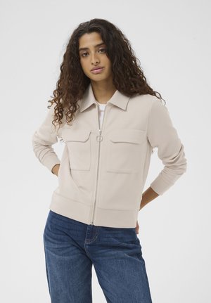 Jonge vrouw met lang krullend haar draagt een beige jas met rits en twee borstzakken, en een blauwe spijkerbroek, staand met haar handen in haar heupen.